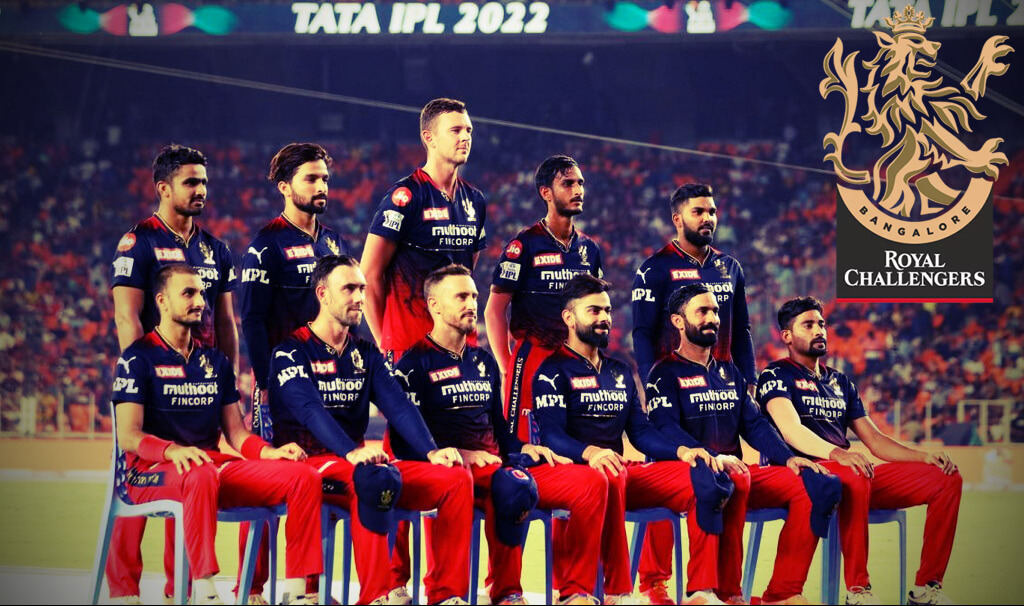 A group of eleven cricket players from the Royal Challengers Bangalore (RCB) team posing for a photo. They are standing in a line, wearing their red and black team uniforms. The players are holding their cricket bats and standing on a green field with a cricket pitch visible in the background.